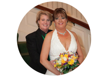Two women standing together, one in a white dress holding flowers, indoors.