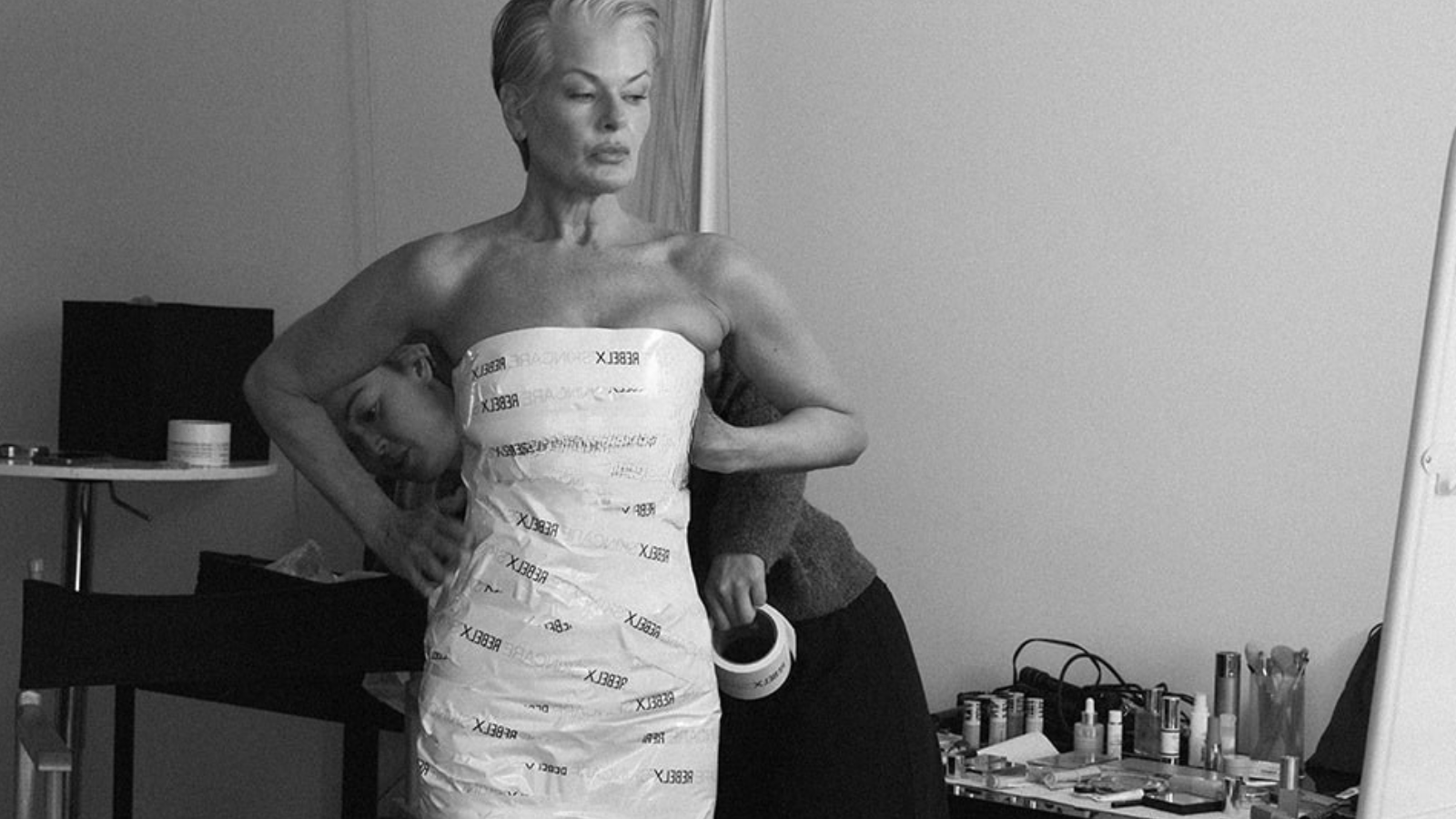 Tina Rowhanian, founder of REBEL X Skincare, wearing strapless white dress with REBEL X logo print in black and white portrait photo with skincare products in background